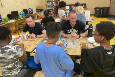 Two male medical students sitting at a table across from three young boys working with building materials.